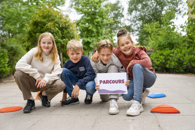 Groep leerlingen kijkt lachend met een bord ‘Parcours’ na de beweegles.
